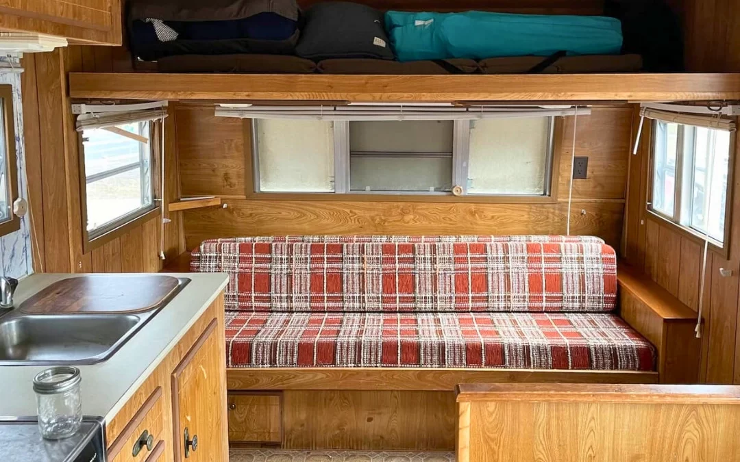 Interior of a small vintage camper showing clever storage hacks with overhead bunk, plaid dinette, and compact kitchen cabinetry