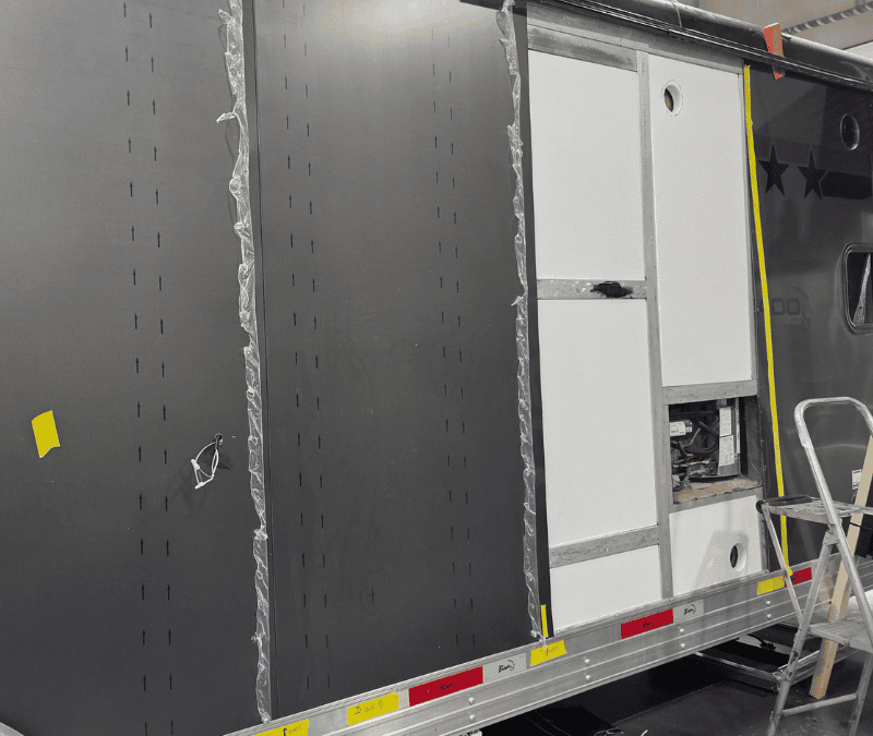 Black perforated metal side panels being installed on a Bison cargo or utility trailer, with white cabinet doors, protective film, and ladder in workshop