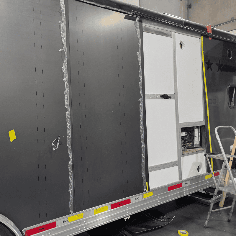 Black perforated metal side panels being installed on a Bison cargo or utility trailer, with white cabinet doors, protective film, and ladder in workshop