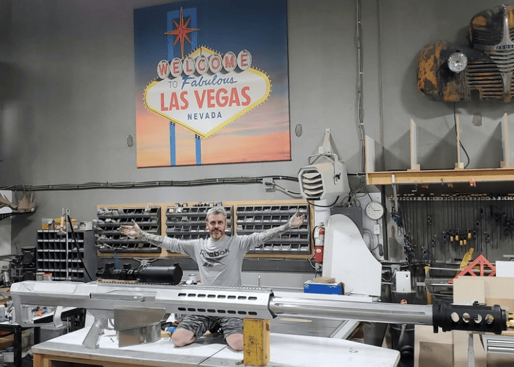 James West of Ace of Spades Custom RV posing with a large custom aluminum-welded Barrett .50 caliber sniper rifle replica smoke stack in his Las Vegas workshop